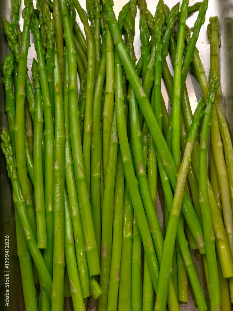 Fototapeta premium A vertical, close-up shot of a bunch of bright green, cooked or blanched asparagus spears lying in a shiny metal tray.