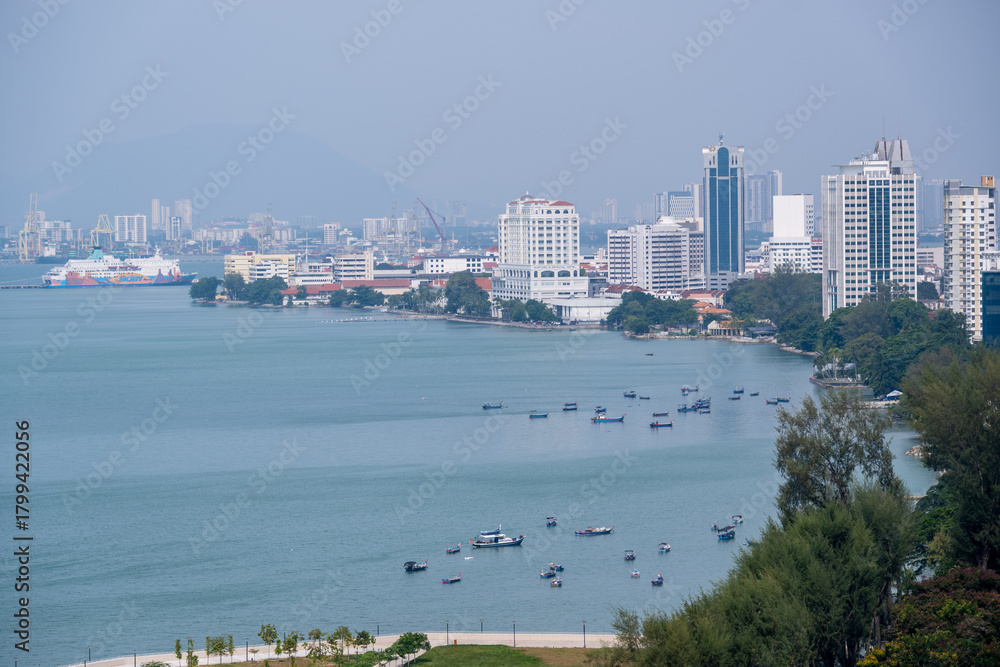 Fototapeta premium Fishing boats rest along the blue waters of Gurney Bay in Penang, framed by modern high-rise buildings and a scenic coastal view.