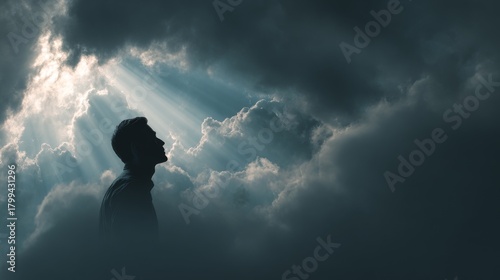 Silhouette of a man looking up, gazing towards divine light breaking through dramatic, dark clouds. A spiritual moment