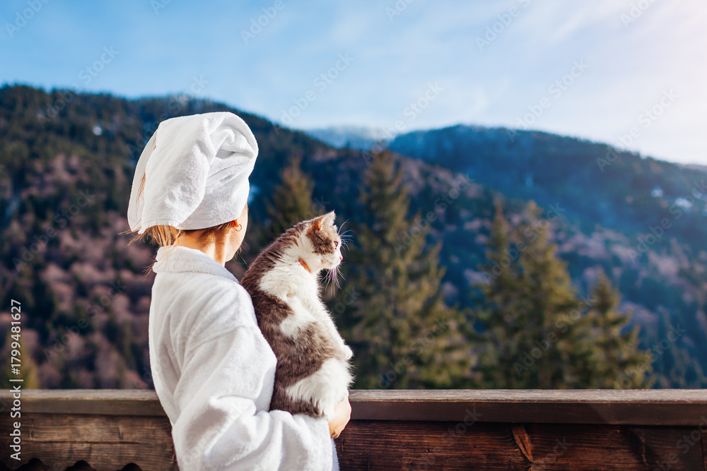 Obraz premium Woman holding cat on hotel balcony enjoying winter mountain landscape wearing bathrobe. Travel with pet