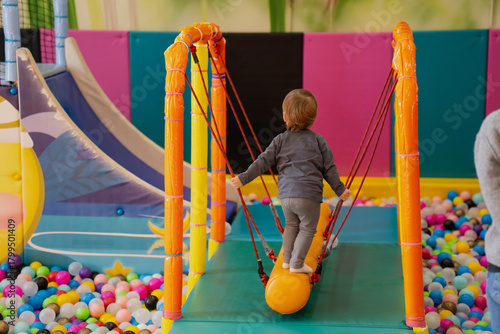 Toddler balancing on a soft cylindrical beam inside a colorful indoor playground, holding support ropes while walking across a padded bridge above a pit filled with plastic play balls