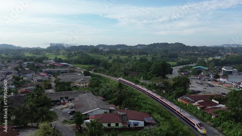 Drone footage captures a train passing through a suburban landscape in Malaysia, showcasing houses, lush greenery, and distant hills under a bright sky.