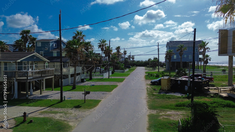 Obraz premium Residential street in Beachside Village, Galveston, Texas features stilt house vacation homes with balconies, palm trees, gravel lot parking, utility poles, ocean views under clear coastal sky