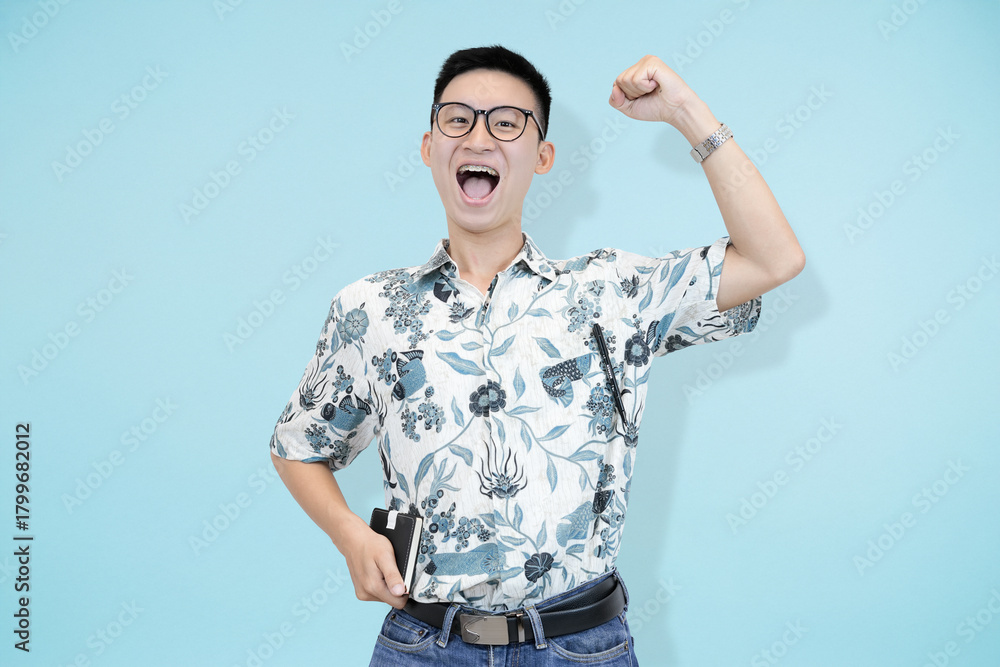 Naklejka premium A cheerful and confident young Asian man and batik shirt, standing confidently in blue studio background