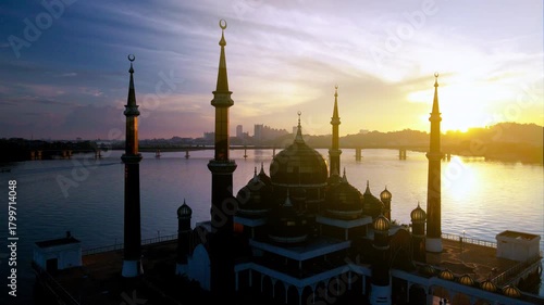 Aerial view of the Crystal Mosque at sunrise, with golden light reflecting on calm waters and clear blue skies around the iconic Islamic landmark.