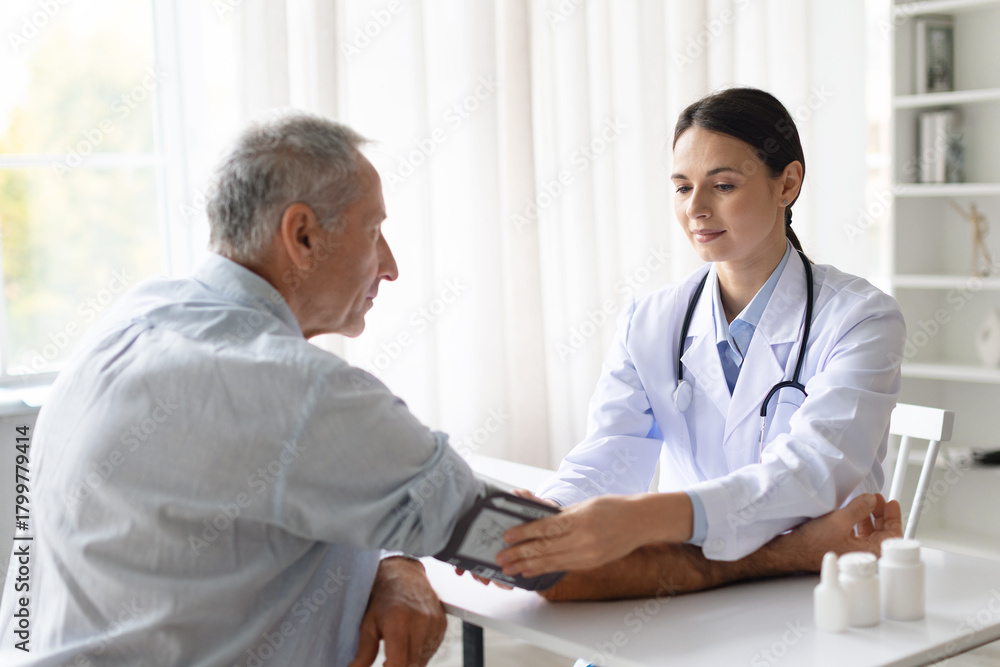 Fototapeta premium Female doctor in white coat measuring blood pressure of senior male patient during medical checkup in clinic, professional healthcare service concept