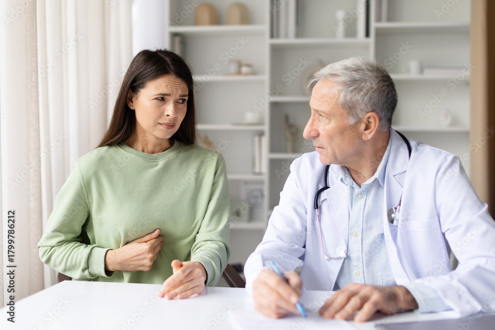 Fototapeta premium Pregnant woman feeling unwell, touching stomach and talking to senior male doctor in clinic office, maternity healthcare and pregnancy care concept