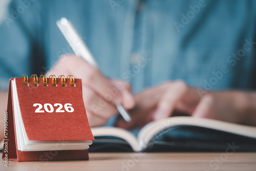 Showing a 2026 desk calendar with a person writing in a notebook representing planning organization future goals time management and preparation for the upcoming year in business and personal life