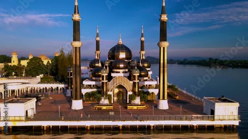 Aerial view of the Crystal Mosque at sunrise, with golden light reflecting on calm waters and clear blue skies around the iconic Islamic landmark.