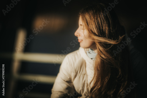 A serene portrait of a young woman with long hair, wearing a white winter jacket, looking away from the camera into the warm sunlight. Her hair catches the light beautifully, creating a soft, dreamy a