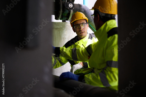 Two engineers in neon jackets and hardhats looking at equipment. A scene of industrial cooperation, captured up close. Focus is on collaboration and attention to detail