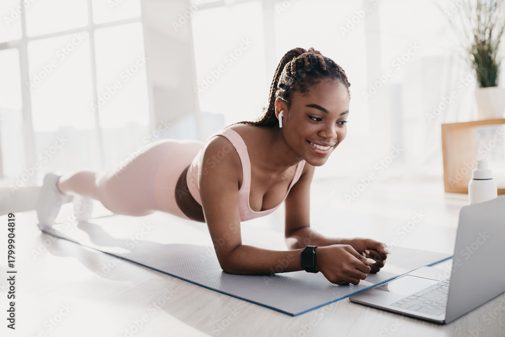 Naklejka premium A sporty millennial black woman performs an elbow plank exercise on a mat in her home gym. She is focused on her laptop with a workout video, smiling as she engages her core muscles.