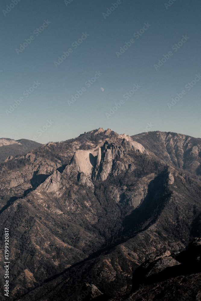 Fototapeta premium A rugged mountain landscape viewed from Moro Rock, with dramatic granite peaks and a faint moon beneath a clear blue sky.