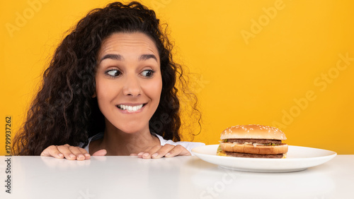 Фотография A playful scene shows a woman peering over a table, her eyes wide with excitement as she looks at a delicious burger on a plate