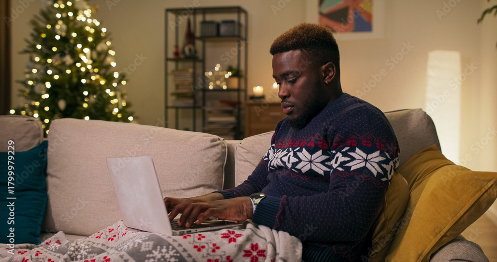 Fototapeta premium Covered with a blanket, a young man of multi-racial background sits on a sofa, typing on his laptop keyboard. He wears a warm sweater, and a Christmas tree is flickering nearby in the living room.