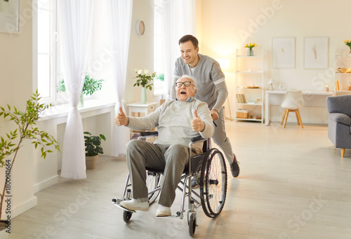 Konstfotografi Joyful senior man with grey hair involved in rehabilitation procedure with young caregiver, using wheelchair for mobility as has walking , having fun in retirement house