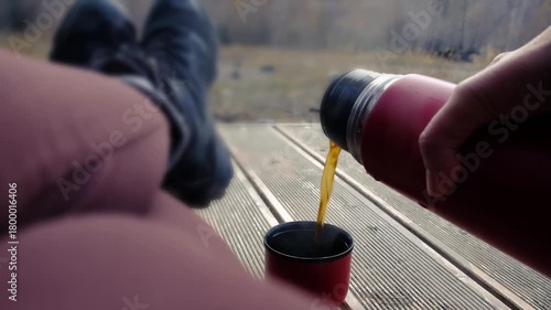 Relaxing outdoor scene, a tourist pours tea from a red thermos, its lid used as a cup, standing on a wooden deck next to a woman