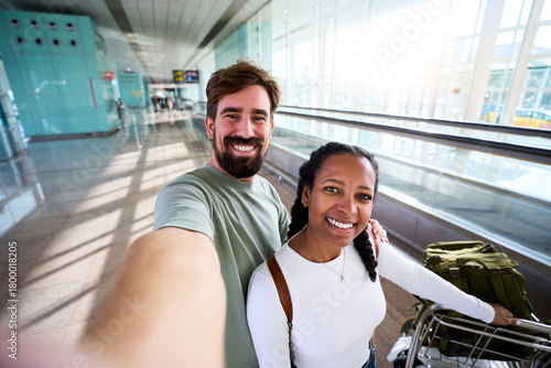 Happy interracial couple taking a selfie at an airport terminal, smiling beside a luggage trolley. Bright glass corridor and walkway suggest boarding, travel excitement and connection.