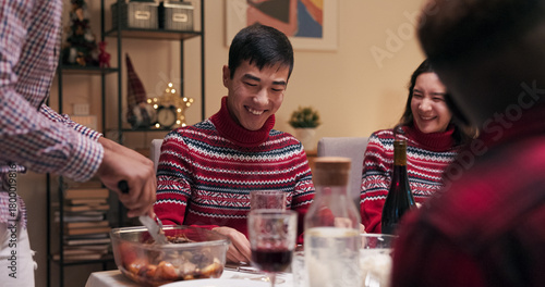 Celebrating the beginning of the holidays, students sit at the table. A girl and boy are visibly dressed in matching red warm sweaters. A guy with dark hair brings up a plate of meat and puts it on