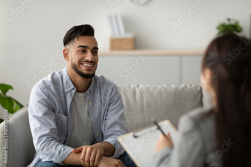 Obraz na płótnie A middle eastern man sits comfortably on a couch in a mental health clinic, smiling as he talks to his psychologist