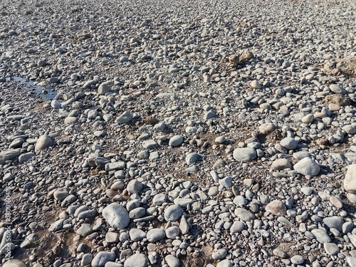 Dry riverbed covered with smooth grey pebbles and rounded stones of different sizes creating a natural rocky texture in bright sunlight.