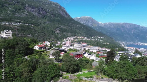 Drone flying over Norwegian fjords, capturing city houses along the shoreline, calm blue water, and steep surrounding mountains in a scenic Nordic landscape.