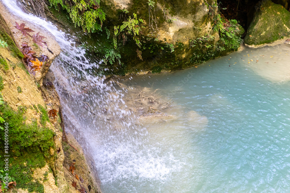 Naklejka premium A small waterfall cascades down a rocky, mossy wall into a natural turquoise pool. Dense vegetation, vines, roots, and moss can be seen all around. (saklı, gizli cennet şelalesi) Manavgat - Antalya