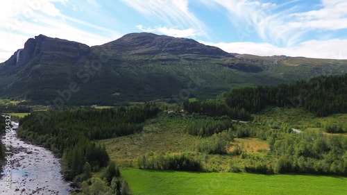 Drone flying over green Norwegian mountains, capturing a waterfall flowing down the hillside and a wide rocky river below in a scenic natural landscape.