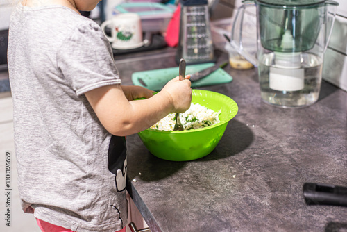 A childs hands actively mix white cottage cheese and green herbs in a green bowl on a dark kitchen countertop