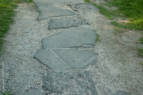 Old broken asphalt path with gravel pushed into the edges, flanked by green grass. Scene of ruins and abandonment, showing wear and neglect.