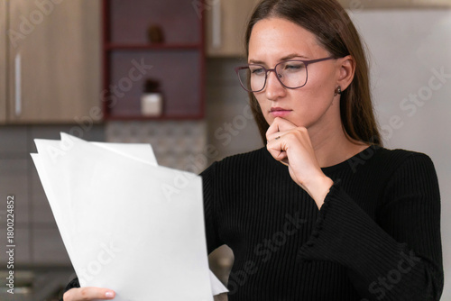 Young woman wearing glasses, reading through important documents while thoughtfully resting her chin on her hand, sitting in her cozy kitchen and reflecting on her financial situation