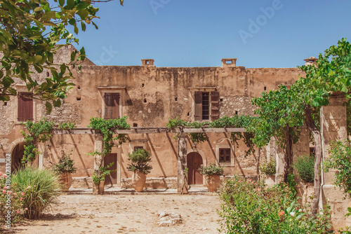Cuadro en lienzo A rustic courtyard building of the Arkadi Monastery in Crete, Greece, featuring weathered stone walls, wooden doors, climbing plants, and warm Mediterranean colors