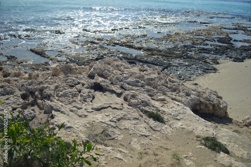 Rocky shore with sandy beach and glistening water under bright sunlight, natural coastal landscape, copy space