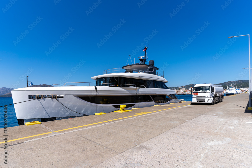 Naklejka premium Luxury motor yacht being refueled the Marmaris marina pier on a clear sunny day, with a fuel tanker truck parked alongside the dock. Modern yacht design, waterfront marina view and harbor background.