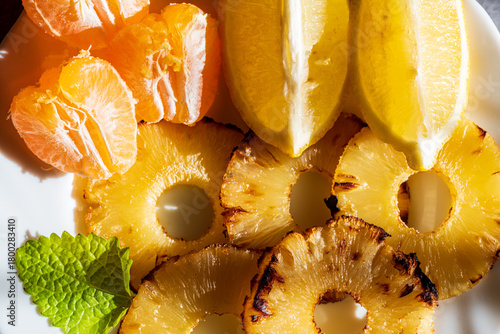 Grilled pineapple slices arranged with fresh mandarin segments and lemon pieces, creating a vibrant and refreshing tropical composition. The caramelized edges of the pineapple add warmth and texture, 