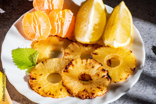 Grilled pineapple slices arranged with fresh mandarin segments and lemon pieces, creating a vibrant and refreshing tropical composition. The caramelized edges of the pineapple add warmth and texture, 