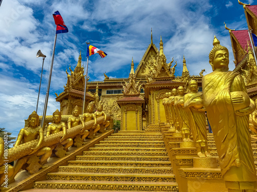 Quadro su tela Golden statues of monks and mythical figures line the grand staircase of an opulent Buddhist temple in Phnom Penh, Cambodia, with national and religious flags flying under a vast, cloud-filled sky
