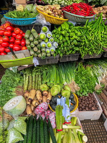 An abundant and colorful assortment of fresh, raw vegetables, including tomatoes, chilies, and lotus root, is artfully arranged for sale at a bustling local market stall in Phnom Penh, Cambodia.