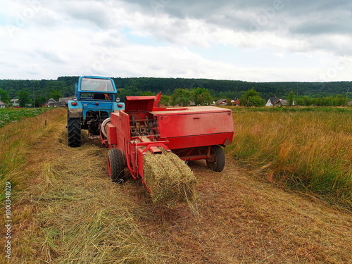 A blue tractor pulls a red hay baler machine, processing cut hay into bales in a rural field