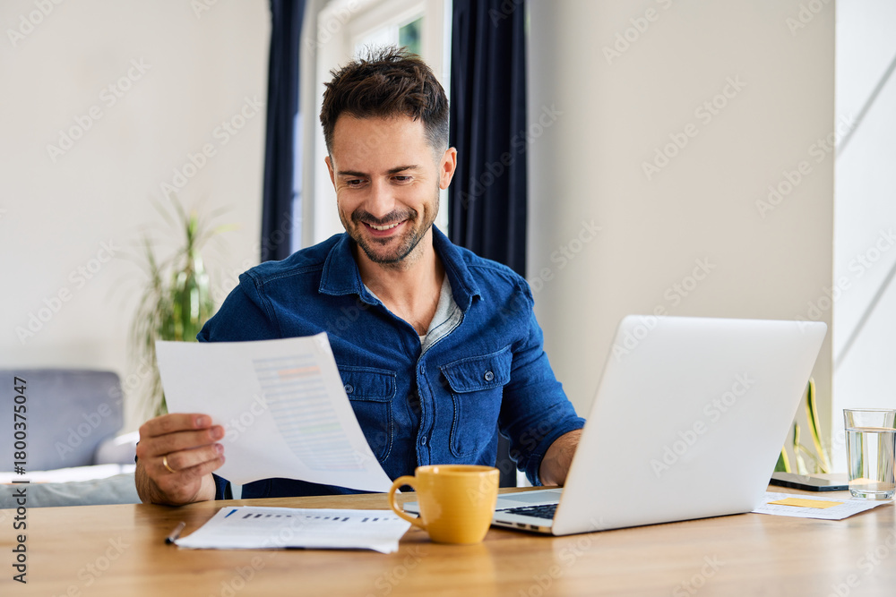 Obraz premium Smiling man looking at financial document working remote on laptop