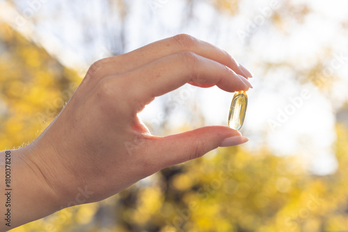 Closeup of a person holding a vitamin D 3 capsule against colourful trees in fall.