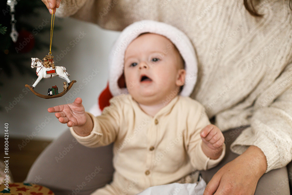 Naklejka premium Happy cute baby in santa hat playing with christmas horse toy with mother under stylish xmas tree. Merry Christmas and Happy Holidays!