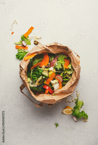 Fresh vegetable peelings and scraps in brown paper bag on light surface, highlighting food waste and its impact