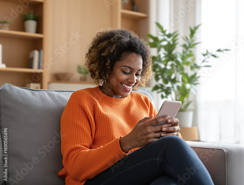 Happy young african american woman smiling while using smartphone on sofa at home