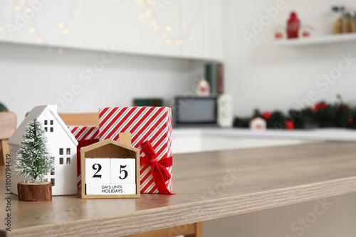 Calendar with text 25 DAYS UNTIL HOLIDAYS, Christmas decor and presents on table in kitchen, closeup