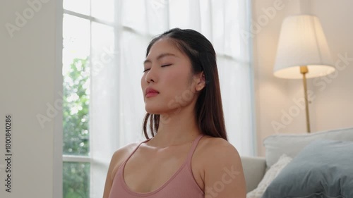 Asian woman in yoga outfit practicing breathing meditation with closed eyes, feeling calm and relaxed at home.