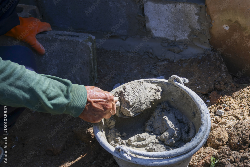Obraz premium Focused manual labor on construction site. person wearing glove mixes gray cement mortar in bucket for foundation repair, showing hard work and skilled effort