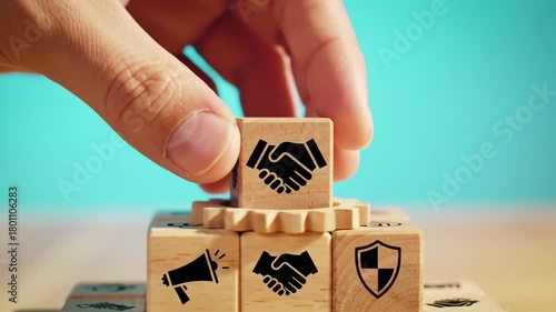 A hand places a wooden block with a handshake symbol atop a stack on a light surface, against a teal background. This arrangement conveys business concepts.
