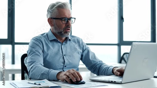 Middle aged gray haired man looking worried while working at a white desk with a computer and office documents in a light filled modern office space.