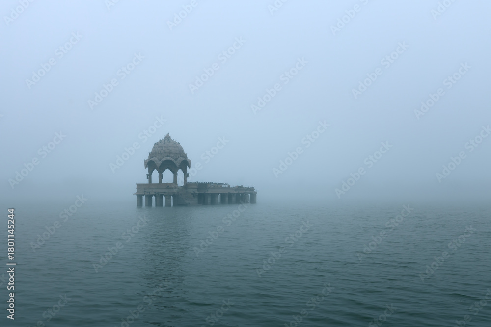 Obraz premium A serene early-morning view of a lone chhatri pavilion rising from the waters of Gadisar Lake in Jaisalmer, Rajasthan, surrounded by soft mist and tranquil reflection.